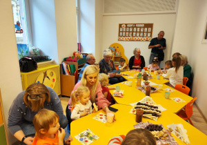 Słodki poczęstunek. Dzieci wraz z dziadkami i babciami siedzą przy stolikach, na których znajdują się ciasteczka.