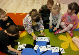 Dzieci siedzą na dywanie. Dziewczynka z drukowanych literek układa wyraz: damy.