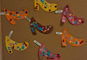 Tablica z pracami dzieci. Na górze znajdują się pantofelki wycięte z kolorowego papieru, ozdobione kolorowymi naklejkami w różnych kształtach. Na dole znajdują się karty pracy z postacią świnki pomalowane kredkami.