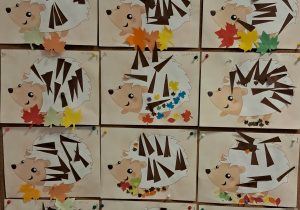 Tablica z pracami dzieci. Rysunki jeżyków wykonane techniką wyklejanki. Każdy jeż ma na grzbiecie przyklejone brązowe trójkąty z papieru, które tworzą jego kolce. Dzieci ozdobiły swoje prace kolorowymi papierowymi liśćmi.