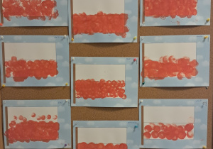 Tablica z pracami dzieci. Rysunki przedstawiające flagę Polski. Dolną część flagi dzieci zamalowały czerwoną farbą, używając do tego stempli.