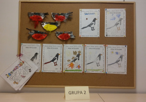 Tablica z pracami dzieci. W lewym górnym rogu tablicy znajdują się wykonane, przy użyciu połowy papierowego talerzyka, piórek oraz farb, ptaszki (gile i sikorka).