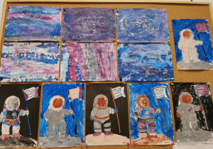 Tablica z pracami dzieci. Na górze znajdują się prace plastyczne przedstawiające Kosmos (wykonane z bibuł, wody i brokatu) Poniżej znajdują się pomalowane farbami postacie misia-astronauty.
