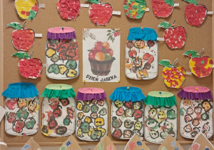 Tablica z pracami dzieci przedstawia prace wykonane z okazji "Dnia Jabłka". Na górze znajdują się kolorowe jabłka wyklejane z bibuły. W centralnej części widoczny jest plakat z koszem jabłek oraz słoiki wypełnione odbitymi farbą mi owocami, ozdobione bibułkowymi wieczkami.