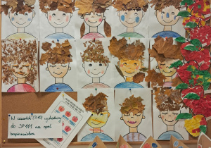 Tablica z pracami dzieci. Postaci dzieci z włosami zrobionymi z jesiennych liści. Po prawej stronie widoczne są wyklejane jabłka z kolorowej bibuły, a na dole – stożki z kolorowymi owocami pomalowanymi kredkami.
