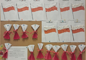 Tablica z pracami dzieci. Na górze znajdują się flagi wyklejone kawałkami białych chusteczek oraz kawałkami czerwonego papieru. Na dole znajdują się czerwono-białe harmonijki przewiązane różowymi wstążkami oraz z przyklejonymi buźkami.