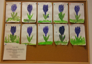 Tablica z pracami dzieci. Na tablicy znajdują się pomalowane farbami, na fioletowo i zielono, kontury kwiatów. Na dole kartki dzieci domalowały trawę.