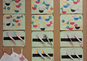 Tablica z pracami dzieci. Na górze znadują się ptaszki wykonane z kolorowych figur geometrycznych. Na dole znajdują się ptaszki pomalowane kredkami z przestrzennymi skrzydłami, siedzące na czarnej gałęzi.