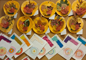 Tablica z pracami dzieci. Na tablicy znajdują się jesienne kompozycje wykonane z pomalowanych farbamina pomarańczowo papierowych talerzyków, ususzonych liści oraz elementów dekoracyjnych. Na dole znadują się prace z wyklejonymi plasteliną literami "L" oraz pomalowanymi kredkami lizakami.