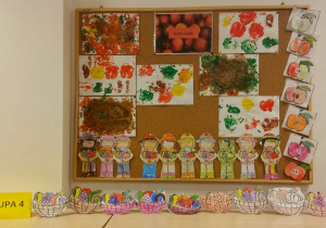 Prace plastyczne dzieci - jesienne oraz z okazji Dnia Jabłka - na korkowej tablicy zostały powieszone: pokolorowane i wycięte postacie ogrodników, wycięte i pokolorowane szablony jabłek, prace wykonane za pomocą stemplowania jabłkami, umoczonym w farbie, kartki papieru. Na szafce stoją wycięte i pokolorowane koszyki z owocami