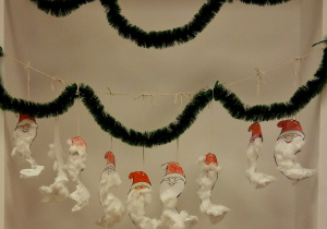 Na zielonym łańcuchu choinkowym (zawieszonym pomiędzy ścianami) wiszą papierowe głowy św. Mikołaja. Czapki są pokolorowane na czerwono, brody są wyklejone watą.