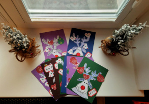 Kartki świąteczne wykonane przez dzieci: wycięta z białego papieru głowa renifera (przyklejona na kolorowy papier), rogi są ozdobione kreskami wykonanymi za pomocą mazaka, nos jest pomalowany na czerwono. Na kartce naklejone się: dzwonki, bombki, serduszka wycięte ze świątecznego papieru.