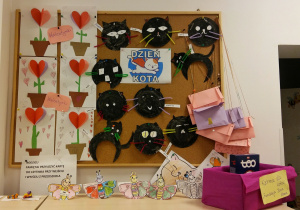 Tablica z pracami dzieci. Po lewej stronie znajdują się kartki walentynkowe wykonane przez dzieci z kolorowego papieru (kwiatek w doniczce w kształcie serca). Po prawej stronie znajdują się koty wykonane z papierowego taleczyka pomalowanego na czarno, wąsy zostały zrobione z drucików kreatywnych, a oczy wycięte z papieru. Na szafce stoją walentynkowe motylki (motylki mają serduszka na skrzydełkach) pokolorowane i wycięte przez dzieci.