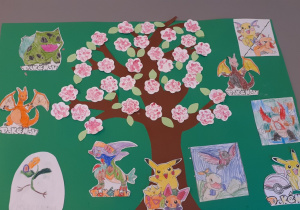 Na stoliku znajduje się brystol, na którym naklejony jest pień drzewa wycięty z papieru oraz białe kwiatki i zielone listki. Dookoła drzewa zostały przyklejone kolorowanki z Pokemonami.