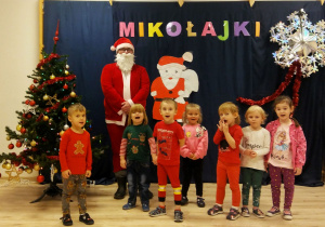 Dzieci z grupy Pszczółek pozują z Mikołajem na tle choinki i napisu Mikołajki.