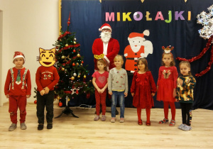 Dzieci z grupy Sówek pozują z Mikołajem na tle choinki i napisu Mikołajki.