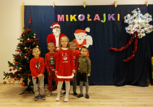 Dzieci z grupy Biedronek pozują z Mikołajem na tle choinki i napisu Mikołajki.