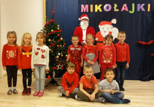 Dzieci z grupy Motylków pozują z Mikołajem na tle choinki i napisu Mikołajki.